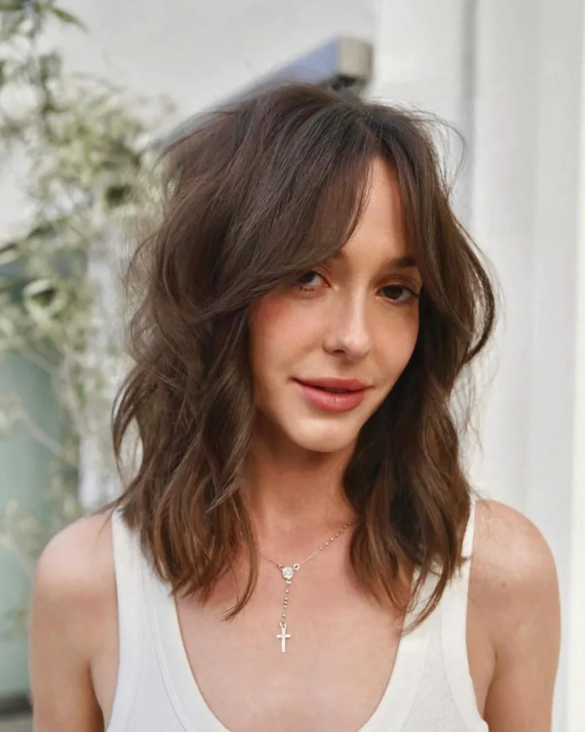 Collarbone-length layered hair with parted curtain bangs that sweep away from the face, warm chestnut color, loose bouncy waves, and a front three-quarter portrait in soft natural window light.