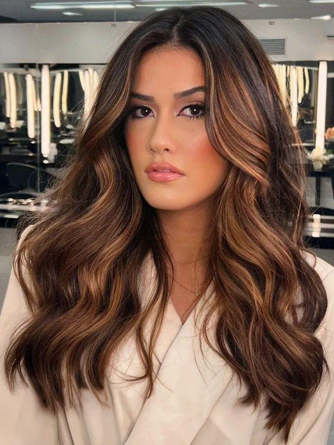 Long naturally black hair with rich medium-caramel ribbons blended through the lower lengths, glossy loose waves, visible contrast without orange tones, controlled soft lighting, and a detailed rear-view salon photograph.