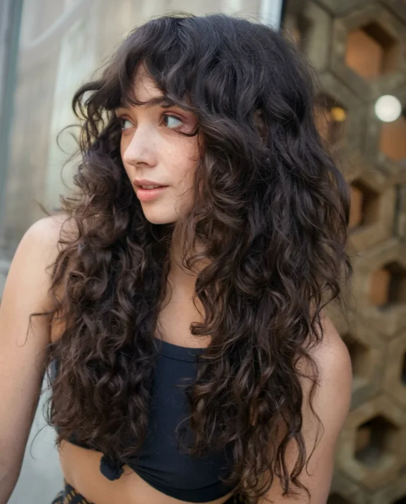 Young woman looking off to the left in a three-quarter view, with very long, dark-brown curls and soft, wispy curly bangs. Layered curls cascade past her chest with lifted volume at the crown. She wears a black cropped top; the background is a blurred stone wall with a honeycomb pattern.