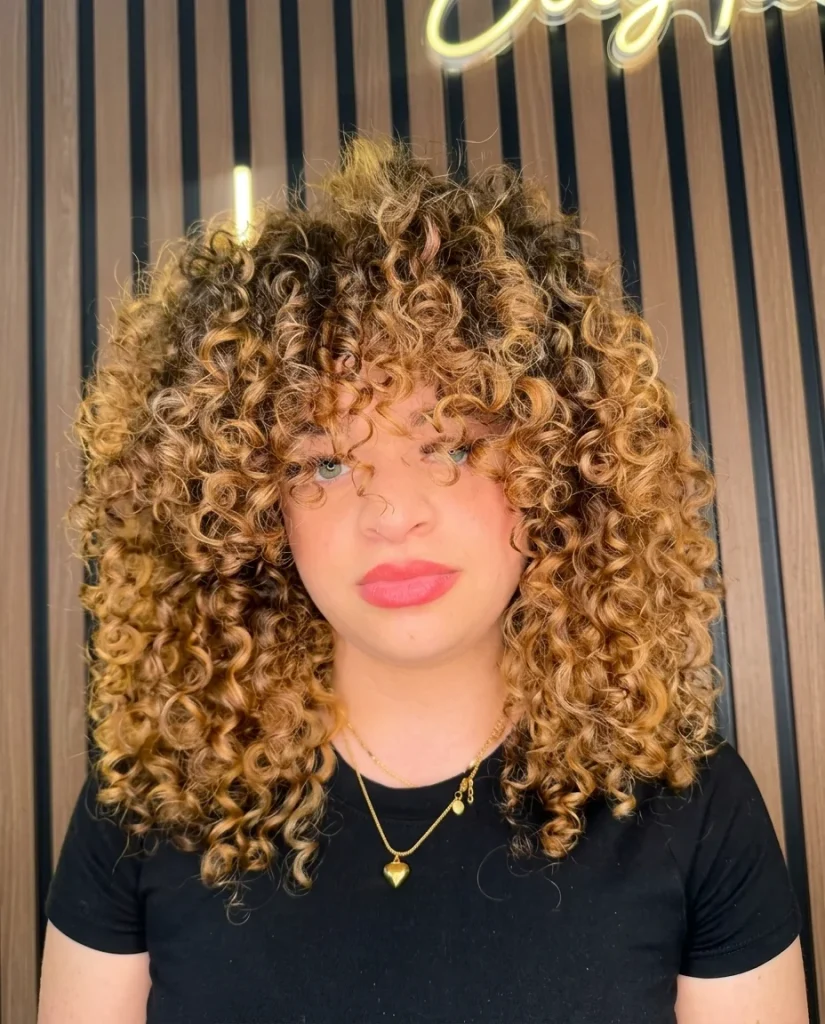 Front view of a young person with medium-length, honey-blonde and caramel highlighted curls shaped into a round, full halo with short curly bangs. They wear red lipstick, a black tee and a gold heart pendant necklace, in front of a wood-slat wall with neon lights. Vertical photo.