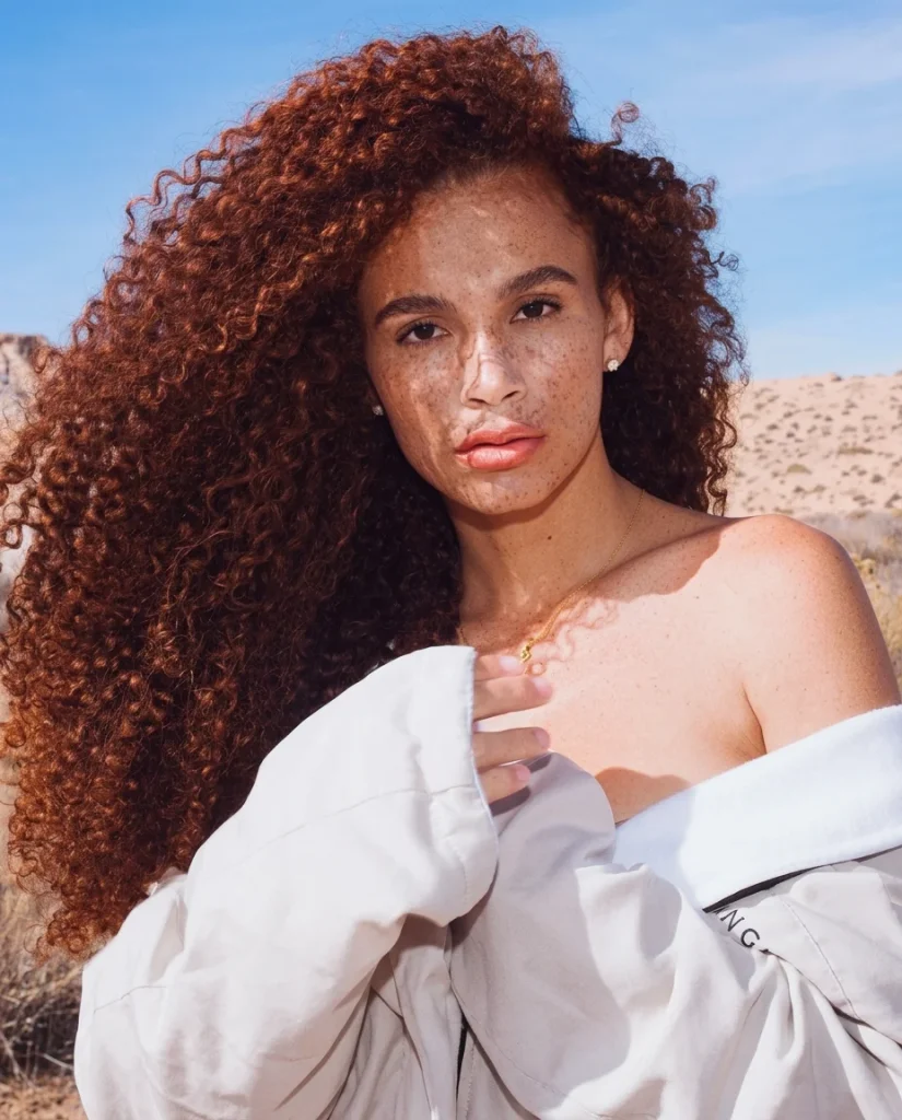Woman with very long, voluminous copper-red curls swept over one shoulder, looking straight into the camera. She has freckled skin, small stud earrings and a delicate gold necklace, and wears an oversized white shirt slipping off one shoulder. Blue sky and desert hills behind her.