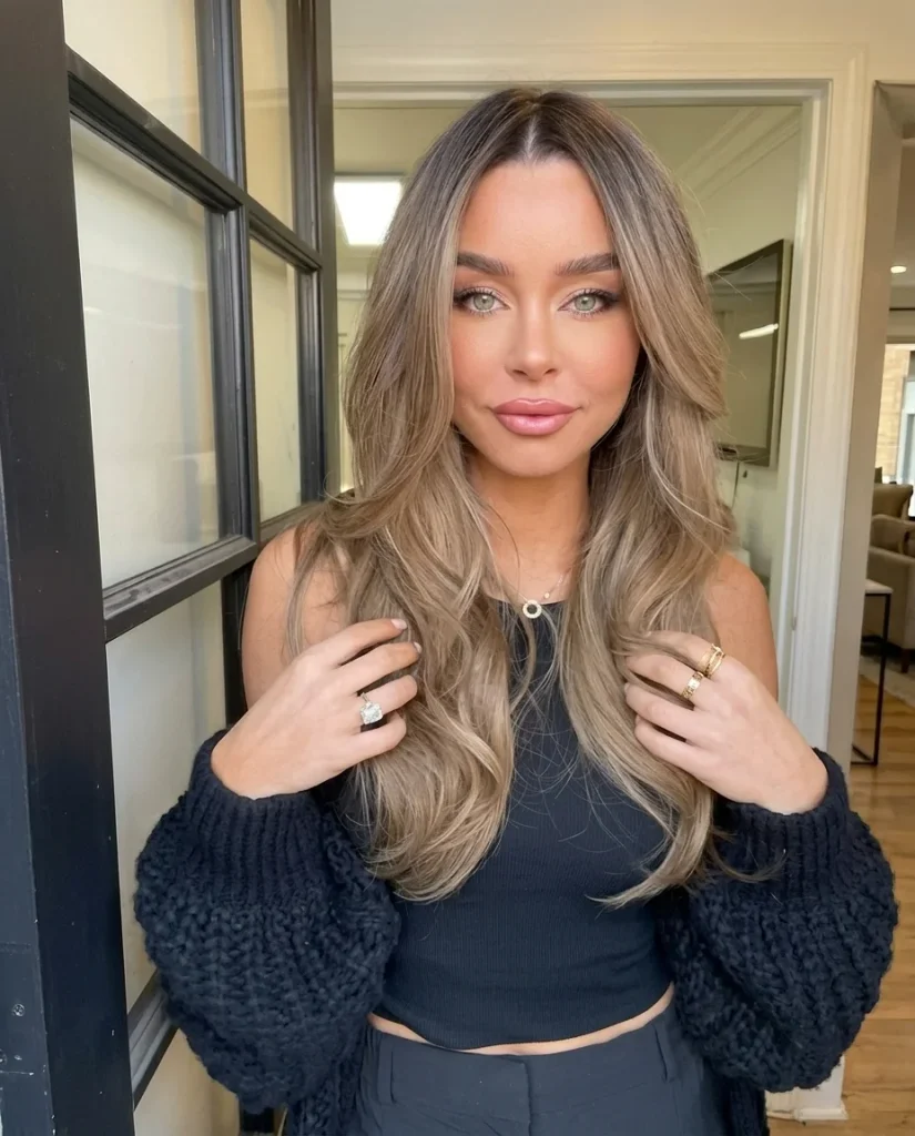 woman with long bronde hair, dark root melting into honey and beige tones, face-framing layers, black knit cardigan, bright doorway