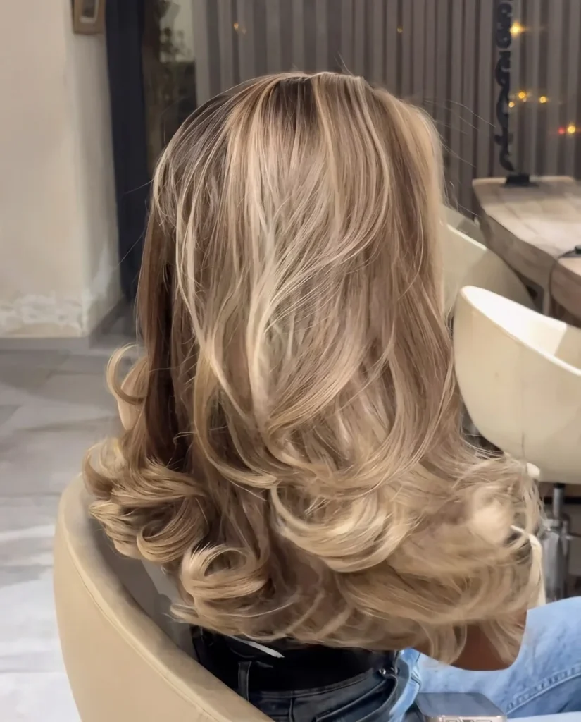 back view of medium-long honey-bronde hair with a dark root and big, bouncy blowout curls, cream salon chair