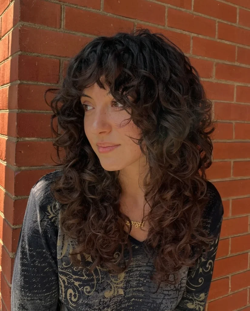 Young woman standing against a red brick wall, glancing to the side with a soft smile. Long, layered dark-brown curls with deep chestnut tones toward the ends, a curly fringe and face-framing layers. She wears a dark long-sleeve top with a gold scroll print and a gold necklace.