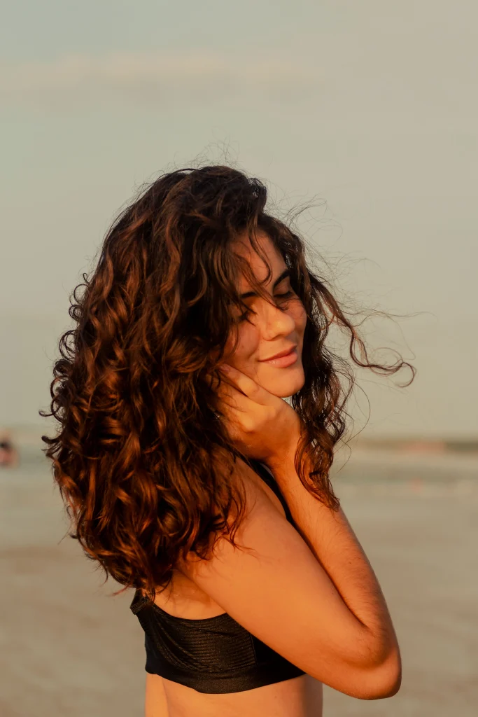 Long natural-looking heatless beach waves, relaxed S-shaped bends, sun-kissed brunette hair, casual center part, soft outdoor daylight, back three-quarter lifestyle photograph.