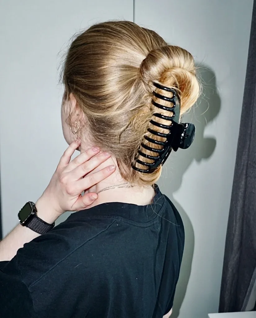 back view of blonde hair twisted up into a bun and held with a large black claw clip, silver hoop earrings, smartwatch, black tee, grey wall