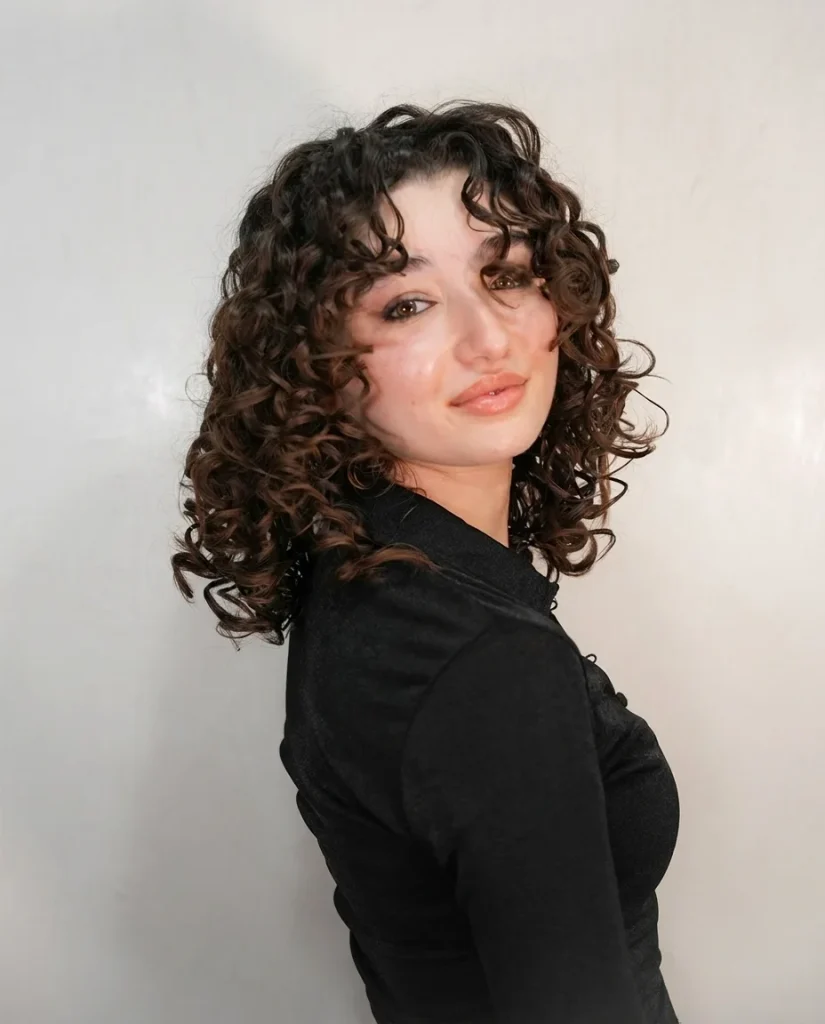 Young woman turned over her shoulder toward the camera with a warm smile. Shoulder-length dark-brown spiral curls with caramel-brown tips, with short curly pieces framing her face. She wears a black collared top against a plain pale wall.