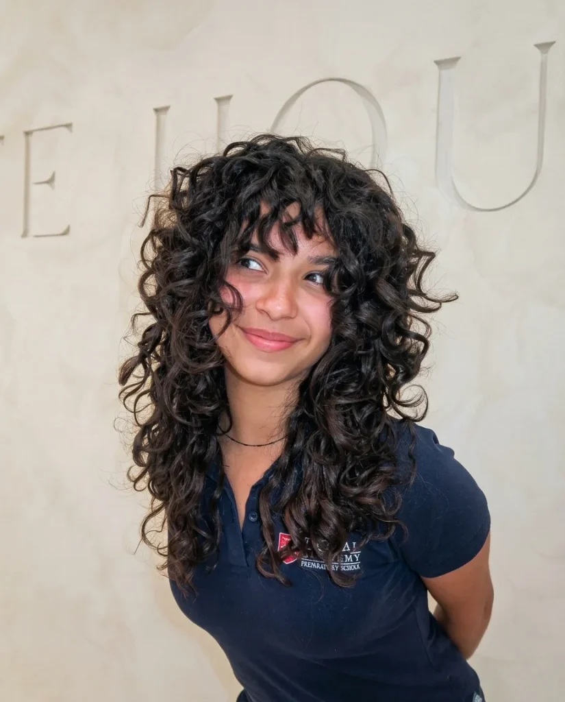 Smiling young person glancing playfully to the side, with long, layered dark-brown curls, a full curly fringe and wispy face-framing pieces. They wear a navy polo shirt with an embroidered school-style logo, standing in front of a warm cream wall with engraved lettering.