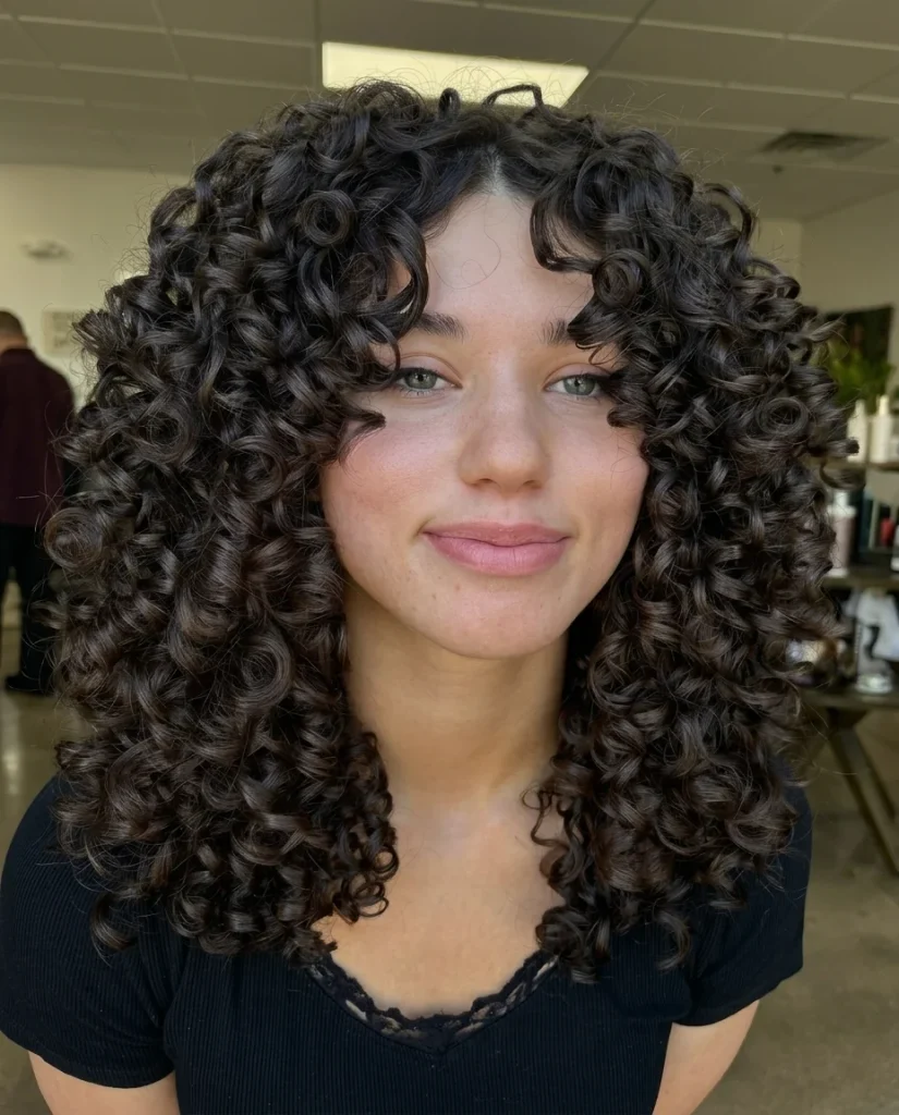 Close-up front view of a woman smiling softly, with a centre part and mid-length, glossy dark-brown spiral curls falling on both sides of her face like curtains. She wears a black short-sleeve top with lace trim, in a bright salon setting.
