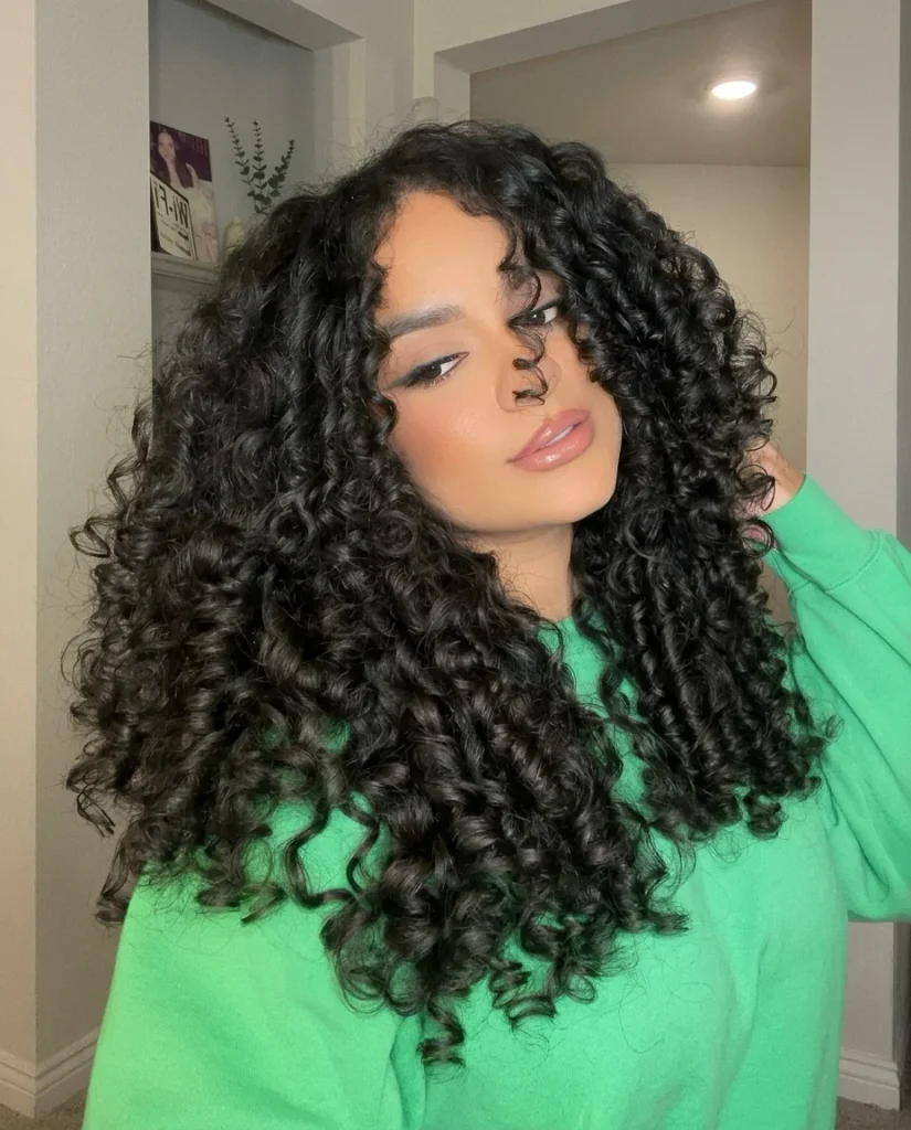 Woman with very long, voluminous black spiral curls, head tilted to one side and one hand in her hair. Glossy, well-defined curls run from roots to ends with side-swept front pieces. She wears a bright green sweatshirt in a neutral indoor setting.