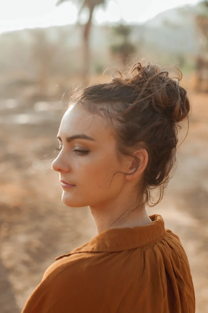 Casual messy high bun with soft face-framing strands, medium-long brunette hair, airy volume and natural texture, bright bedroom daylight, lifestyle beauty portrait.