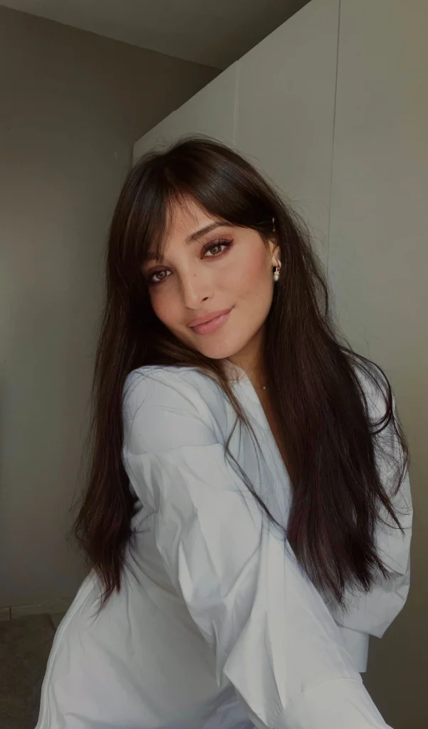 Woman with long, soft brunette hair, wispy curtain bangs parted at the center, feathered cheekbone-length pieces, natural movement, and a flattering front-facing salon portrait.