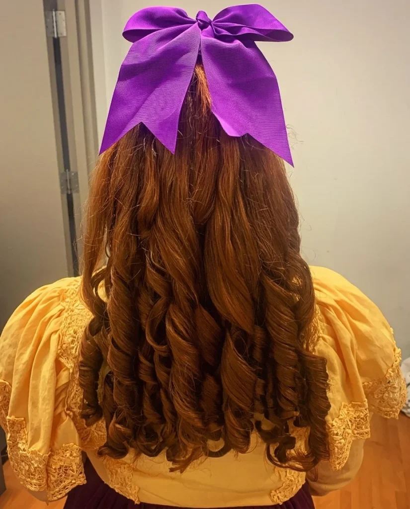 Back view of a person with long copper-auburn ringlets reaching the middle of the back, topped with a large purple bow at the crown. The glossy corkscrew spirals are uniform and well separated. They wear a yellow, lace-trimmed puff-sleeve blouse against a plain indoor wall.
