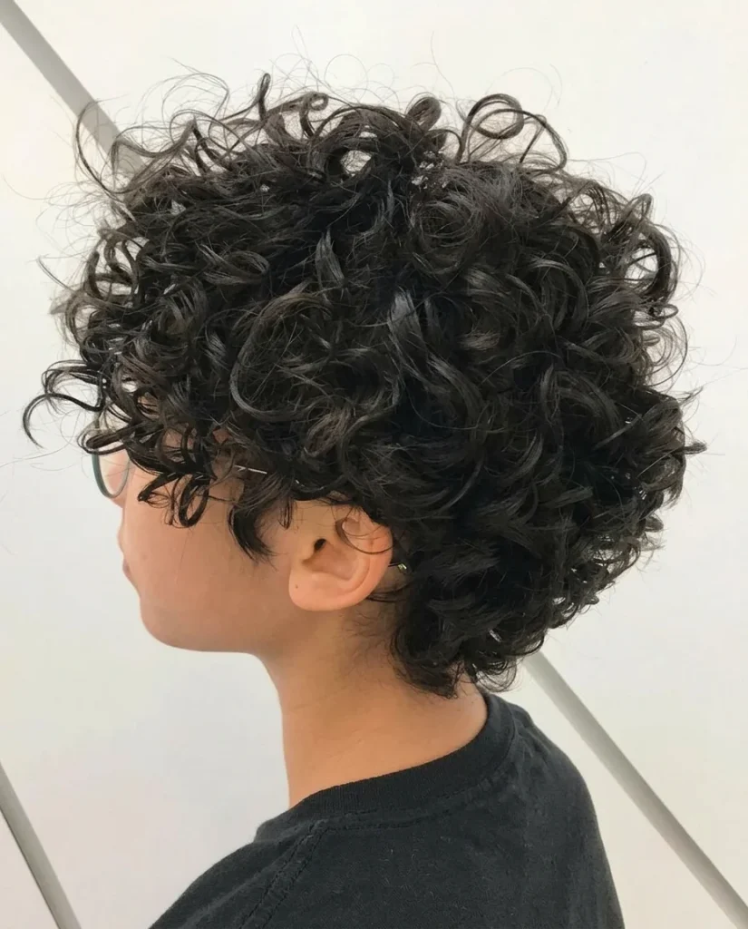 Side-profile view of a young person with a short black curly pixie: close-cropped, tapered sides and nape, and a big, voluminous mass of loose curls on top. They wear a black tee against a white background with grey diagonal lines.