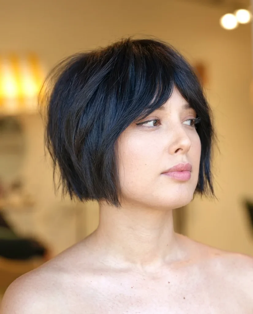 woman in side profile with a blue-black choppy layered bob and soft side-swept bangs, bare shoulders, warm blurred salon lights