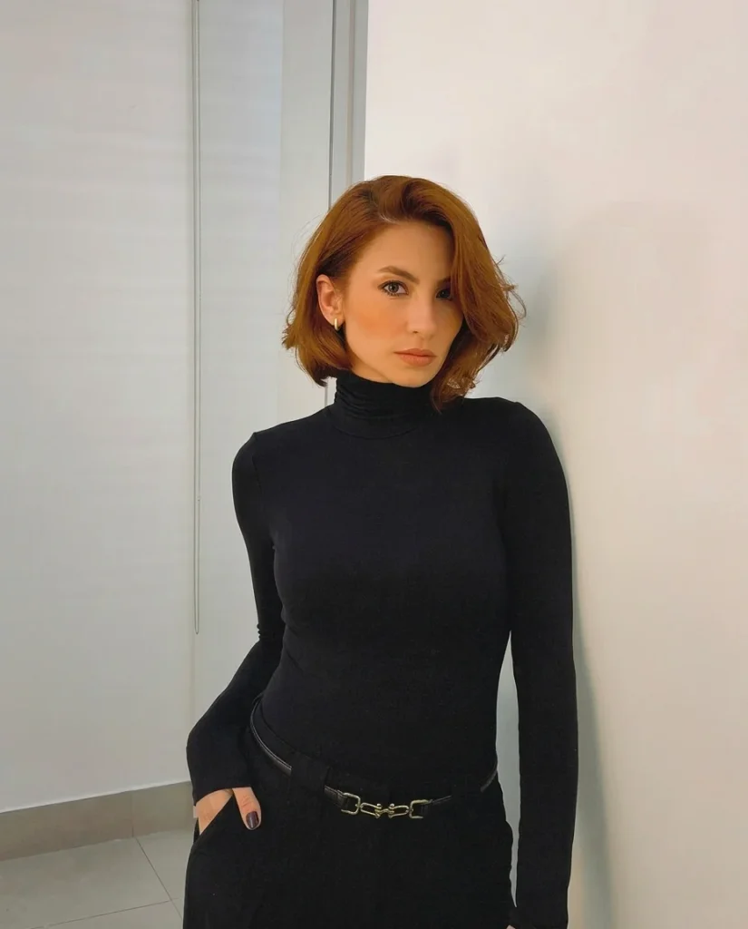 woman with a chin-length copper-red wavy bob and soft side part, black turtleneck and belted trousers, standing against a light wall