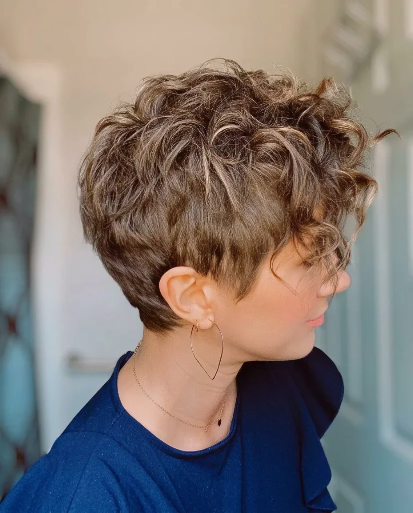 side profile of a woman with a short curly pixie, tapered sides and caramel highlights through brown curls, blue top, gold hoop earring, soft pastel background