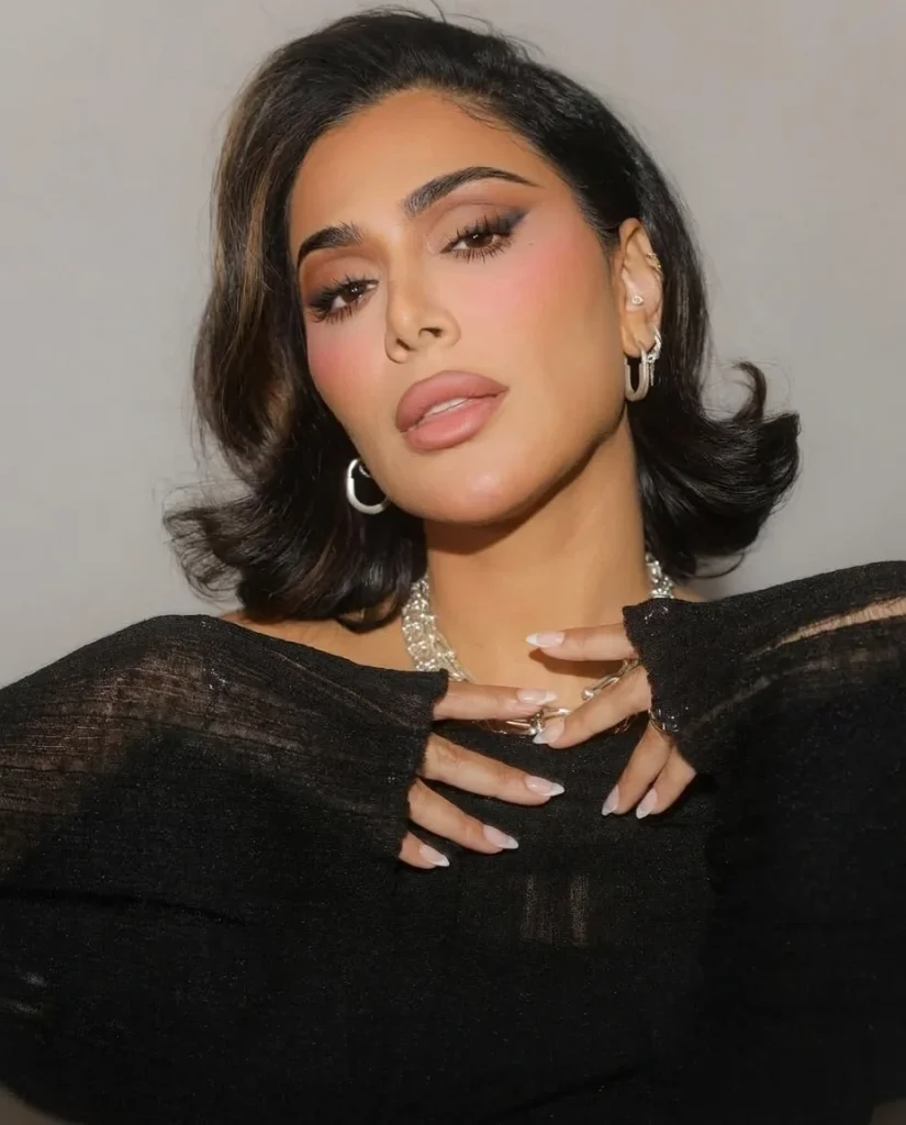 woman with a glossy black bob with golden-brown face-framing ribbons and flipped-out ends, smoky eye makeup, silver jewelry, black sheer knit top, neutral grey backdrop