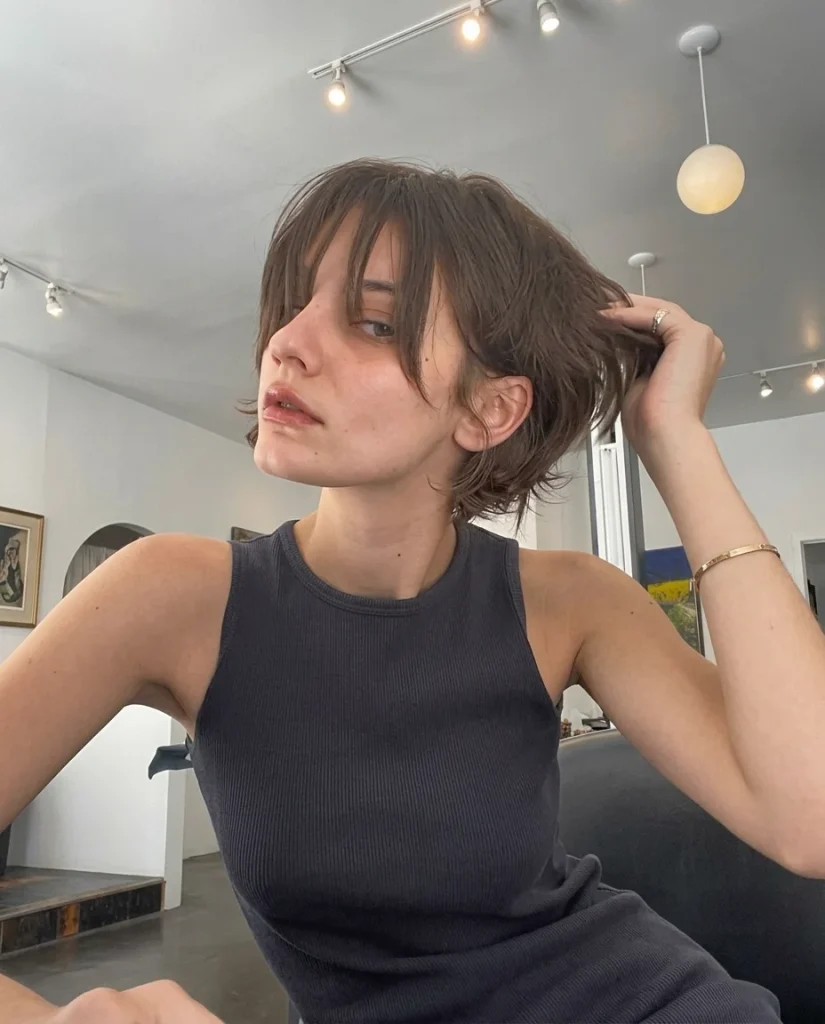 woman with a shaggy brown pixie, wispy eyebrow-grazing bangs and longer layered pieces at the ears and neck, running a hand through her hair, charcoal tank top, bright gallery-style interior