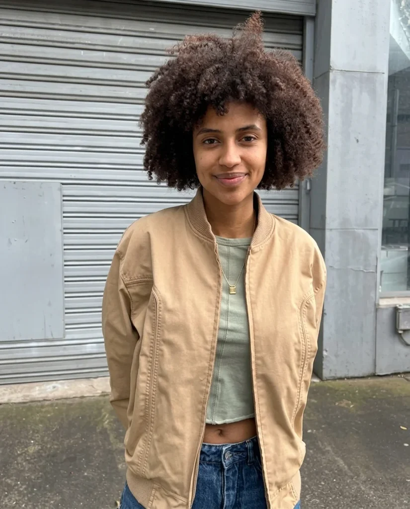 smiling woman with a short, rounded natural curly cut with tight springy coils and copper-brown tips, tan bomber jacket, sage green top, grey street background