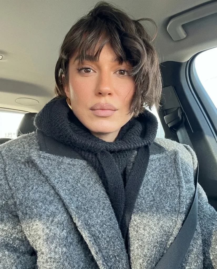 woman with a short dark-brown tousled French bob, wavy undone texture and wispy bangs, gold earring, grey wool coat and black knit hood, car interior selfie
